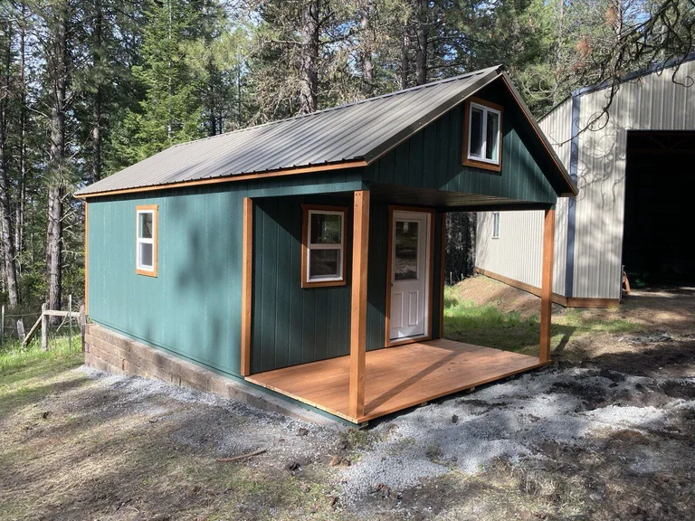 Green cabin with metal roof, wood porch deck, and loft window above the entry