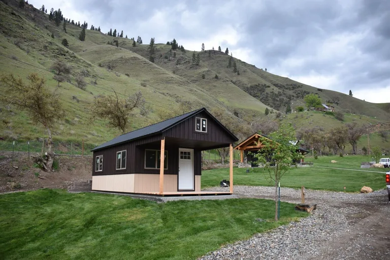 Dark brown two-tone cabin with white door and porch posts below grassy hills