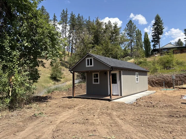 Gray cabin with black shingle roof and covered porch on a freshly graded homesite