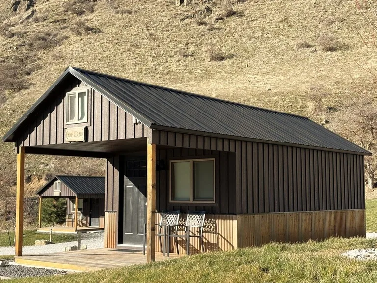 Dark brown cabin with metal roof, large windows, and porch chairs at the base of a canyon