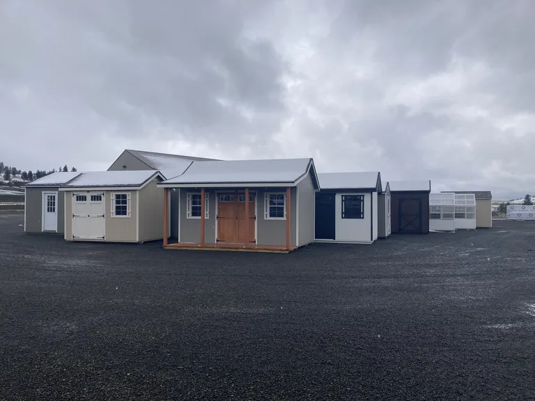 Gray carriage style shed with wood double doors, transom windows, and covered porch on a display lot