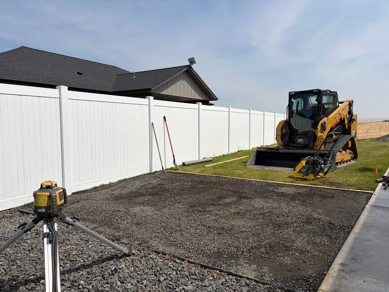 Prepared gravel shed pad beside a white fence with a laser level and compact track loader