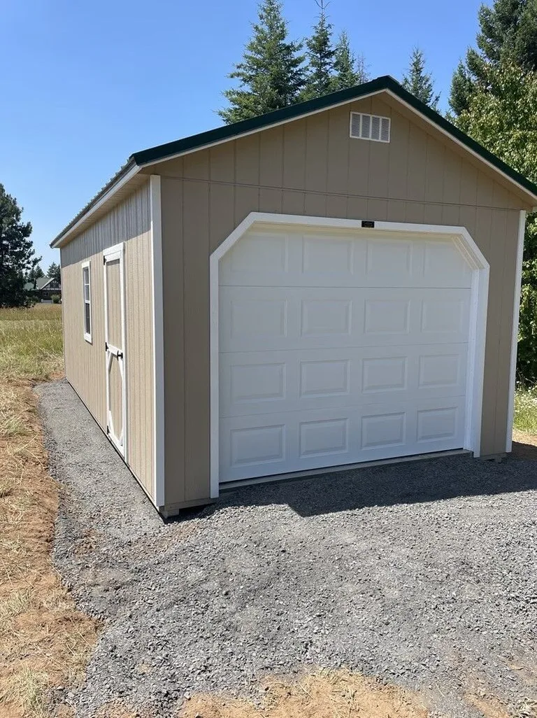 Tan single-car garage with white overhead door and green roof on a gravel pad