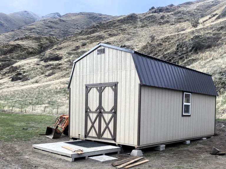 Tan high barn shed with a gambrel metal roof and double doors beside an Idaho hillside