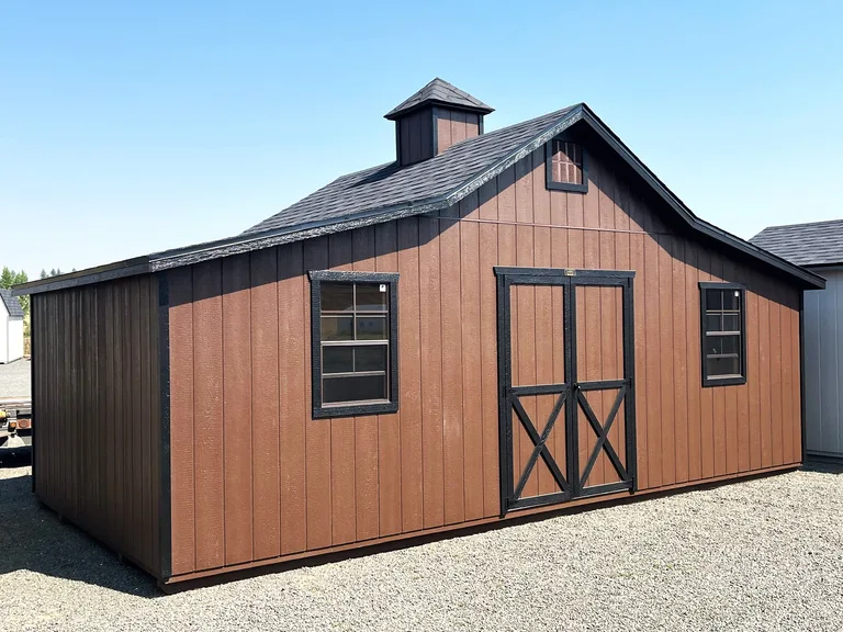 Brown Rancher barn with black trim, cupola, double doors, and gable loft window on a gravel lot
