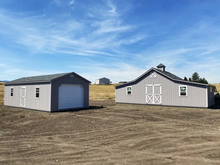 Light gray Rancher barn with cupola and white trim next to a matching garage on open ground