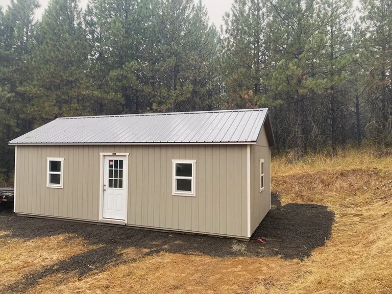 Tan utility style shed with metal roof, entry door, and windows